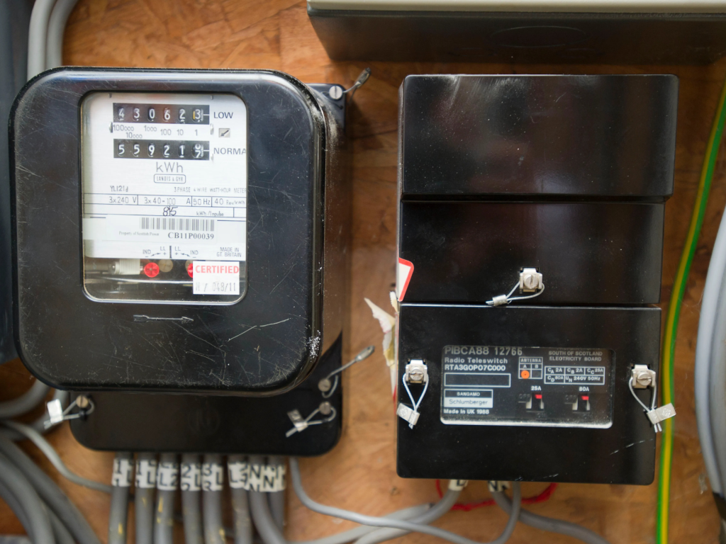 A close-up photograph of an electricity meter and a radio teleswitch mounted on a wooden board. The meter has a black casing with a glass display showing kilowatt-hour readings, certification labels, and wiring connections at the bottom. The adjacent black teleswitch box has labeled switches and a security seal. Multiple electrical cables are connected to both devices.